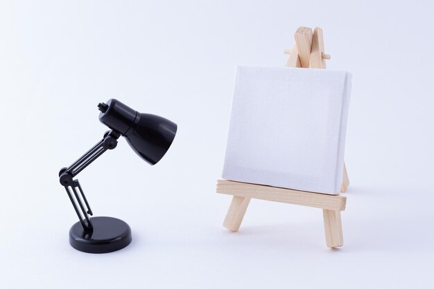 アーティスト​や​画家​の​ため​の​空白​の​白い​正方形​の​キャンバス​と​木製​イーゼルミニチュアモックアップ​きれいな​アート​ボード​と​白い​背景​の​コピー​スペース​に​小さな​黒い​テーブル​ランプ付き​の​ミニ​木製​スタンド