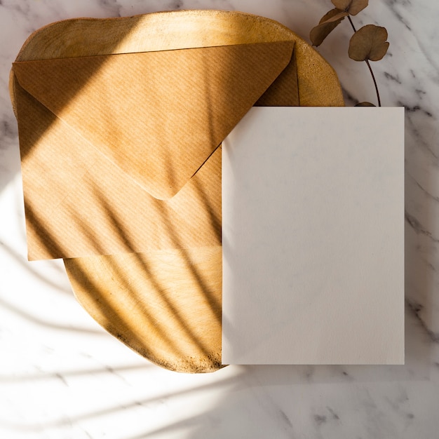 Wooden log and leaf branch with a brown envelope and a white blank on a marble background with leaf shadows