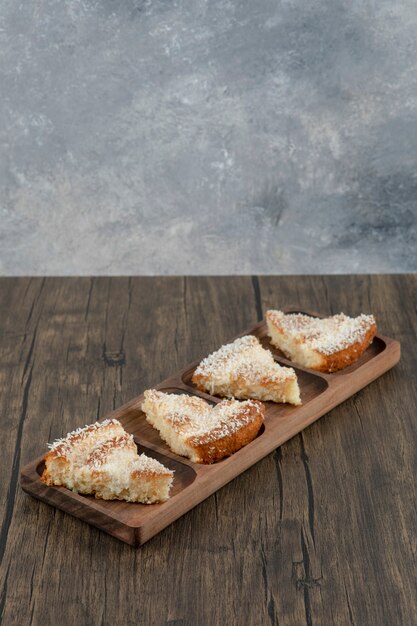 Wooden plate of delicious cake slices with coconut sprinkles on wooden table.