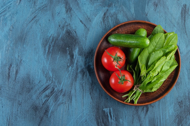 Wooden plate of fresh tomatoes, cucumbers and greens on blue background. 