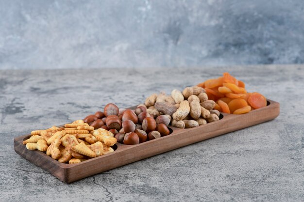 Wooden plate full of various nuts, crackers and dried apricots on marble surface.