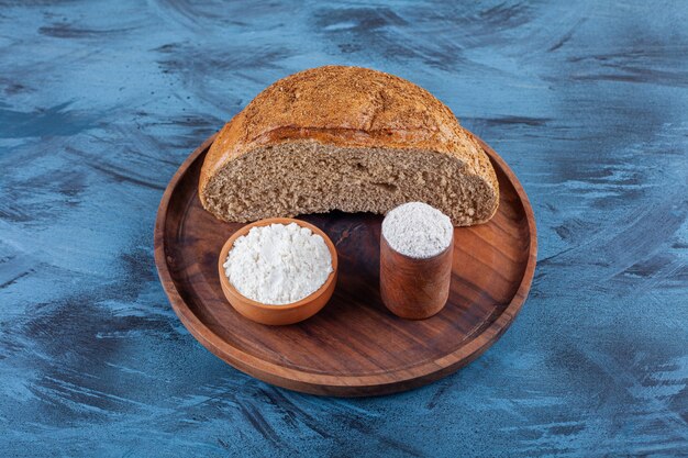 Wooden plate of halved rye bread with bowl of flour on blue surface.