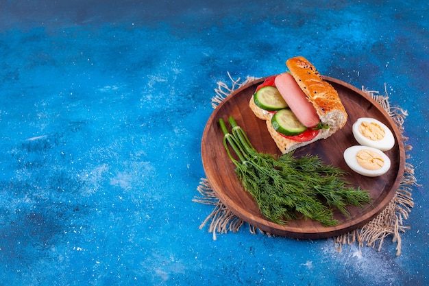 Wooden plate of sausage sandwich with dill and eggs on blue surface. 