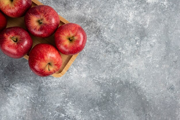 Wooden plate of shiny red apples on marble table.