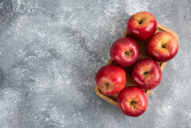 Wooden plate of shiny red apples on marble table.