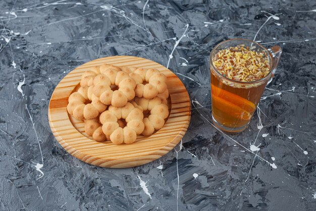 Wooden plate of sweet flower shaped cookies and cup of tea on marble surface.