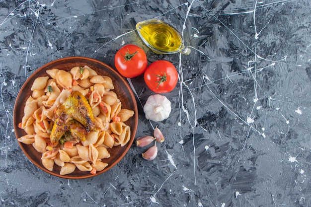 Wooden plate of tasty seashell pasta with chicken wing on marble surface. 