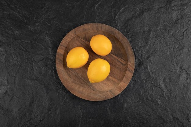Wooden plate of three fresh lemons on black table.
