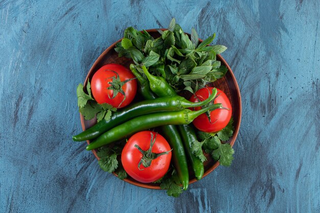 Wooden plate of tomatoes, chili peppers and greens on blue surface. 