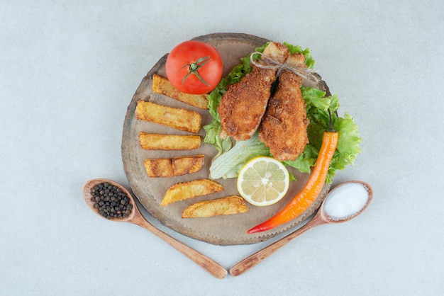 A wooden plate with fried cgicken and vegetables on marble table.