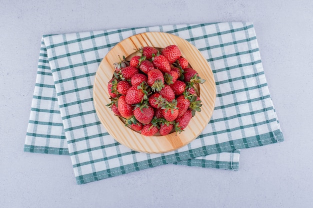 Wooden platter on a folded tablecloth with a pile of strawberries on marble background.
