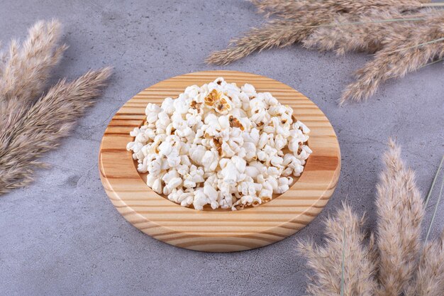 Wooden platter with a portion of popcorn placed in the midst of dried needlegrass on marble background. High quality photo