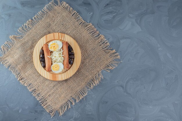 소시지, 얇게 썬 계란, 강판 치즈, 대리석 테이블에있는 직물 조각에 블랙 올리브 나무 플래터.