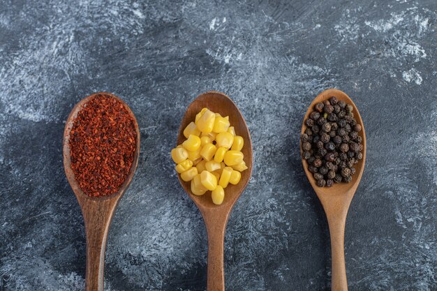 Wooden spoons of corn kernels, ground and grain peppers on marble surface.