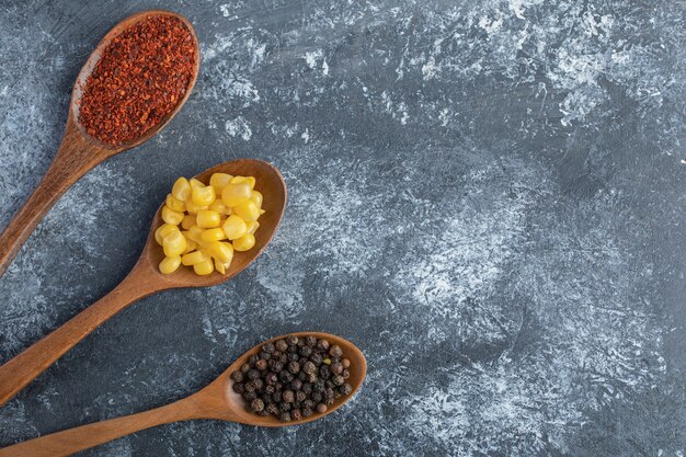 Wooden spoons of corn kernels, ground and grain peppers on marble surface.