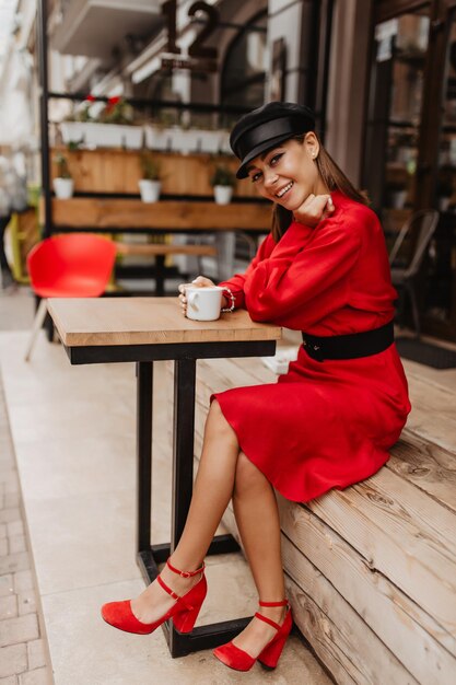 On wooden terrace near cafe young and beautiful girl dressed in bright red dress and red shoes drinks aromatic coffee and looks into camera