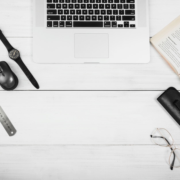 An wooden white table with laptop; mouse; eyeglasses; book and wrist watch on white desk