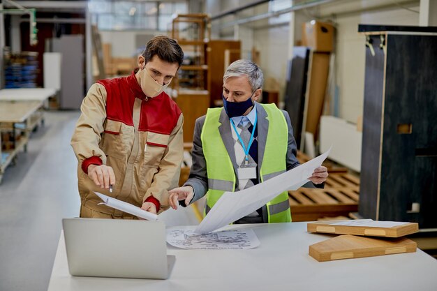 Woodworking engineer and male worker using laptop while analyzing design plans in a factory