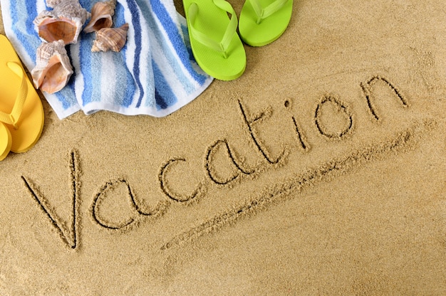 The word vacation written in sand with flip flops and beach towel 