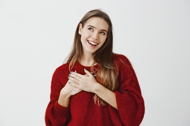 Words hit right in heart. Dreamy beauitful girlfriend in red loose sweater, holding hands on chest and looking up with pleased romantic expression, smiling broadly while imaging positive things