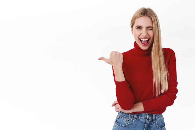 Work career and lifestyle concept Cheerful attractive blonde girl with sassy attitude pointing thumb left and smiling wink encourage you to participate event click banner or download app