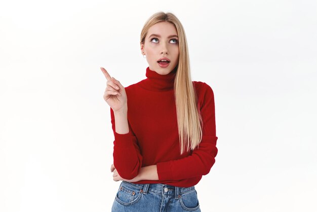 Work career and lifestyle concept Sassy and coquettish blonde woman in red turtleneck pointing finger upper left corner and looking away showing way to promo recommend beauty product