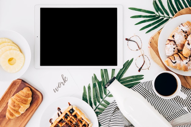 Free photo work text on paper near the digital tablet; pineapple slices; croissant; waffles; bottle; coffee cup and eyeglasses on white desk