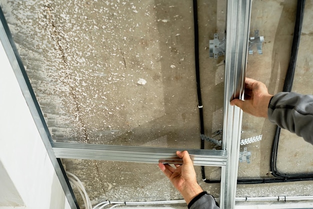 作業員は、石膏ボードの天井フレームを取り付けるための金属プロファイルを調整し、専門家の手にクローズアップして選択的に焦点を合わせます。産業改修と改修