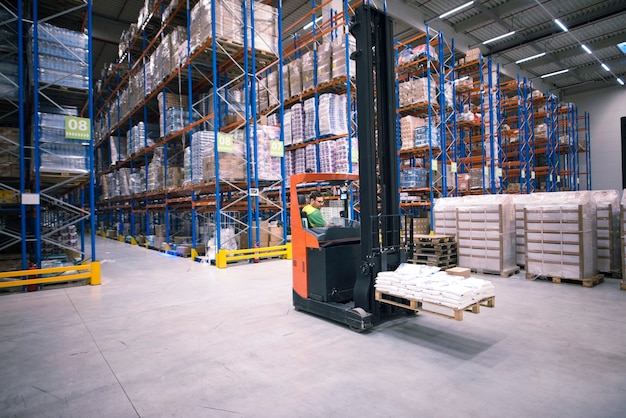 Worker operating forklift machine and relocating goods in large warehouse center