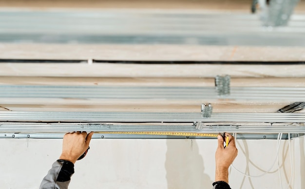 Руки рабочего замеряют и корректируют металлический профиль для монтажа каркаса потолка из гипсокартона, крупный план, выборочная фокусировка в руках специалиста. Промышленный ремонт и реновация