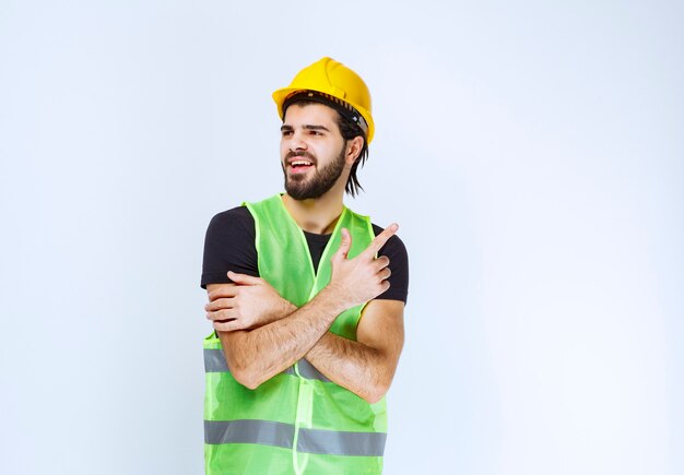 Worker in yellow helmet and green jacket pointing up.