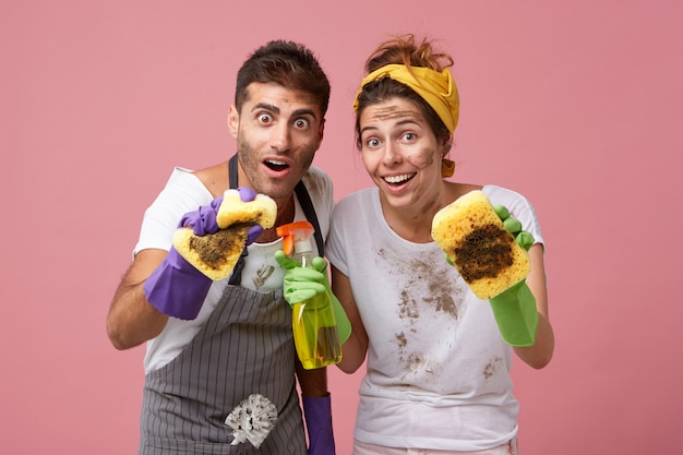 Работники службы уборки вытирают пыль губками. счастливая домохозяйка убирает дом с моющим средством и ее муж с удивленным выражением лица