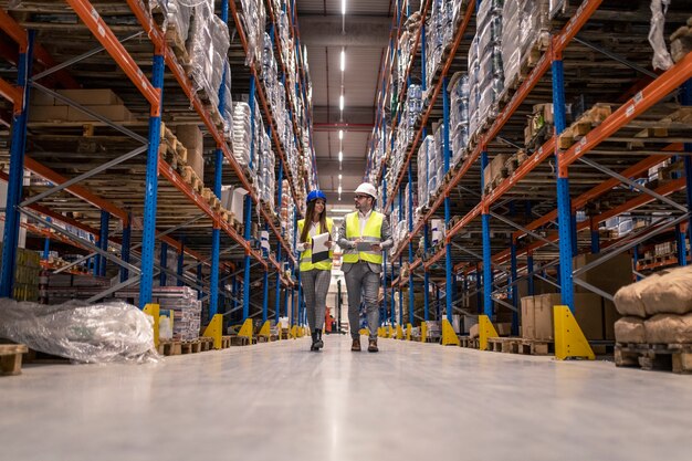 Workers with hardhats and reflective jackets walking through big warehouse aisle checking goods condition