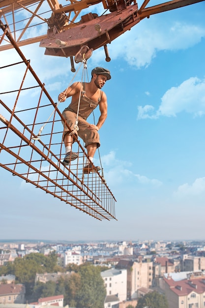 Working carefully. Shirtless strong retro builder looking at something attentively standing on a crossbar hanging from a crane low angle shot