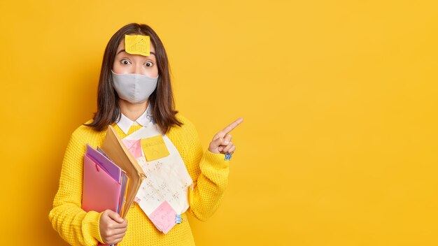 Working during coronavirus pandemic. Surprised Asain woman office worker wears protective mask stuck with papers and sticky notes looks surprisingly indicates at copy space