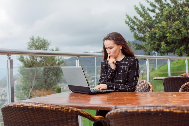 Работающая на ноутбуке девушка-блогер думает, держа указательный палец на подбородке на фоне природы