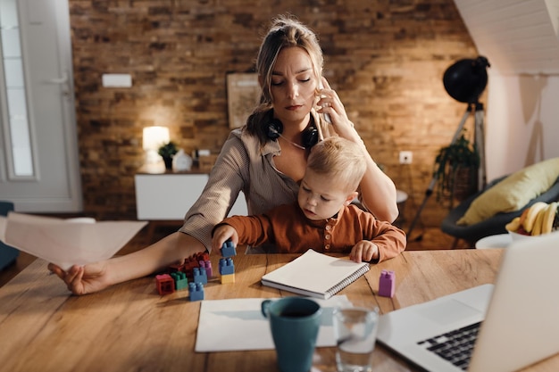 Working mother talking on the phone while her son is playing with toy block at home