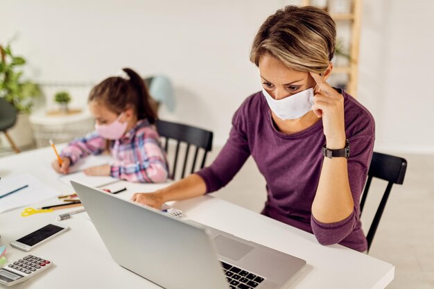 Working mother using laptop while her daughter is doing homework during coronavirus epidemic