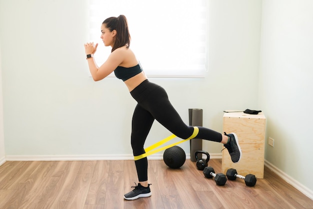 Тренируйтесь, чтобы иметь более сильные ноги. Спортивная женщина тренирует мышцы ног с помощью ленты сопротивления во время кросс-тренировки дома