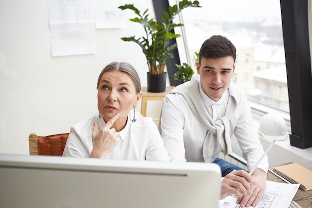 Working in team. Creative architects young man and senior woman developing construction plan in white office, sitting at desk in front of computer, having thoughtful expressions, brainstorming