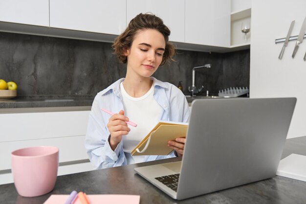 働く女性はメモを作り、自宅でラップトップを持って座っているノートに情報を書き留めます