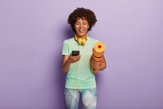 Workout and fitness concept. Cheerful dark skinned woman holds mobile phone connected to headphones