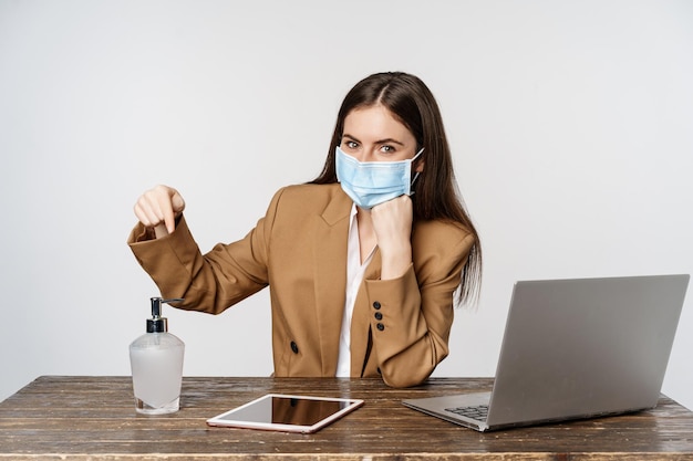 Workplace and covid-19 concept. Portrait of business woman in medical face mask, sitting at table and pointing finger at hand sanitizer, standing over white background