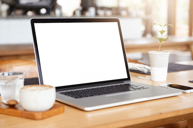 Workplace of self-employed person: generic laptop pc resting on wooden table with smart phone, mug of coffee and glass of water.