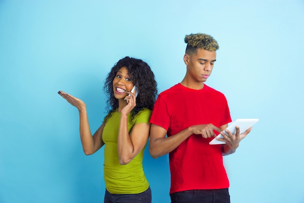 Works on tablet, talks on phone. Young emotional african-american man and woman in colorful clothes on blue background. Beautiful couple. Concept of human emotions, facial expession, relations, ad.