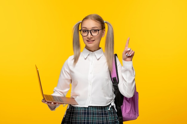 Scolara bionda eccitata giornata mondiale del libro in uniforme con zaino rosa che tiene un computer