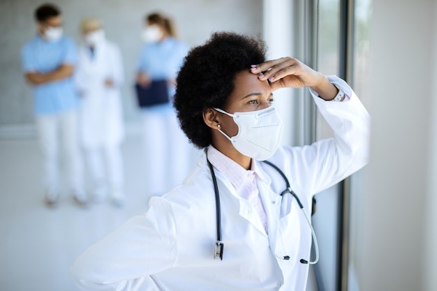 Preoccupata dottoressa afroamericana con maschera facciale che guarda attraverso la finestra