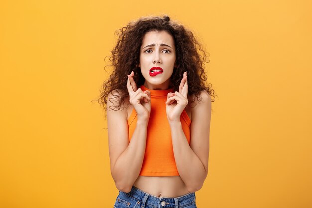 Worried and anxious silly caucasian female with curly hair in red lipstick and cropped top biting lower lip nervously looking concerned crossing fingers for good luck making wish over orange wall.