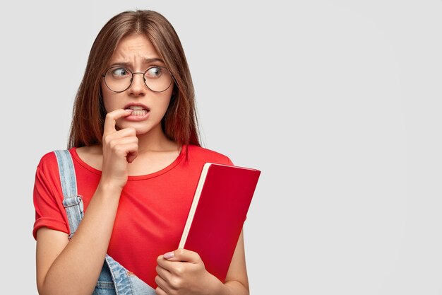 걱정스러운 젊은 여성은 검지 손가락을 입 근처에두고 필사적으로 옆으로 쳐다 보며 둥근 안경을 쓰고 빨간색 교과서를 들고 흰색 벽에 모델을 들고 승진을위한 여유 공간을 제공합니다.