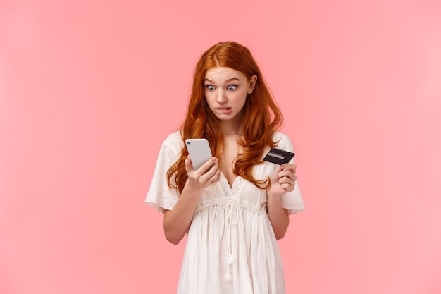 Worried and awkward cute redhead girl made mistake, accidently wasted all boyfriend money during shopping, looking guilty with oops face staring smartphone display, holding credit card